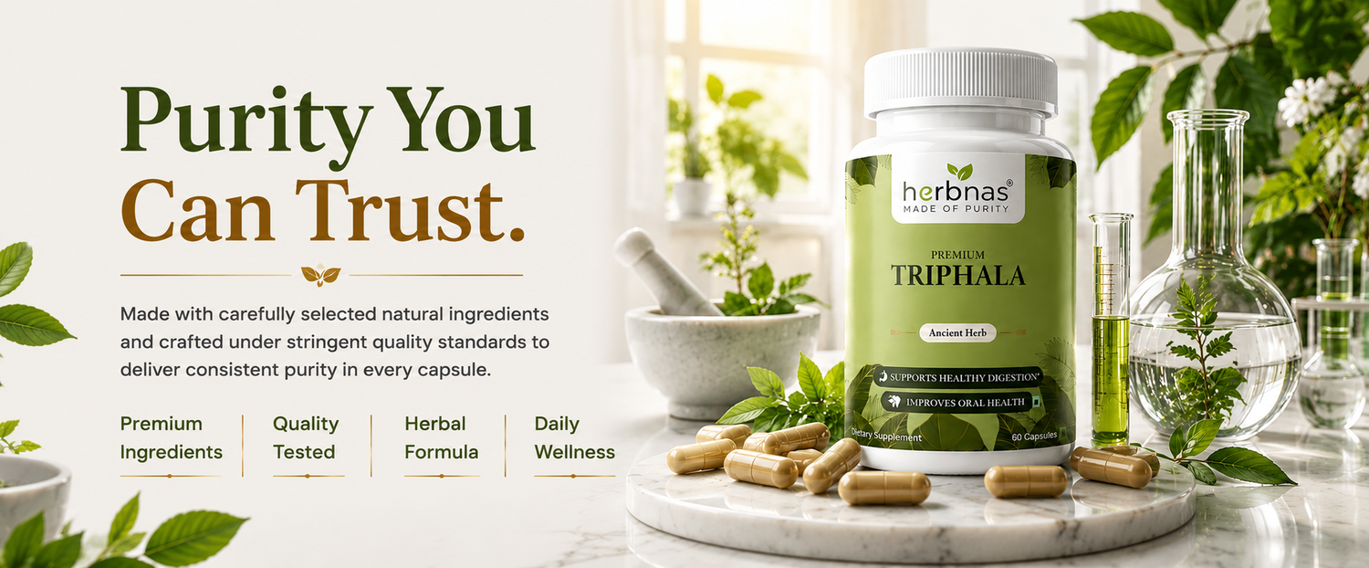 A bottle of herbnas Triphala herbal supplement with capsules scattered on a marble tray, surrounded by green leaves, glass beakers, and a mortar and pestle in a bright, clean studio setting with text on the left.